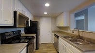 This single-story space showcases a well-organized kitchen as its central feature. The kitchen is designed in a galley-style layout, with cabinetry lining both sides to maximize storage and efficiency. White upper and lower cabinets with sleek hardware provide ample storage, while neutral-toned countertops offer generous workspace for meal preparation. Recessed ceiling lights illuminate the kitchen evenly, creating a bright and practical environment. Appliances include a black refrigerator, a black electric range with coil burners, an over-the-range microwave, a dishwasher, and a double-basin stainless steel sink with a modern faucet. Light wood-look flooring runs throughout, adding warmth and continuity to the space. A white interior door at the far end of the kitchen leads to an adjacent area, contributing to the overall flow of the layout. The clean, neutral color palette of white cabinetry and warm flooring creates a cohesive and functional aesthetic, making the kitchen a practical and inviting area suited for everyday use.