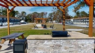 Located at 1834 Wilmington Hwy in Jacksonville, North Carolina, this two-story multi-unit property presents a classic motel-style architectural layout with a white exterior and brown roofline. The building features open-air exterior corridors running the full length of both floors, with railings providing access to individual units on the upper level. Stairwells are positioned at key points along the structure for convenient upper-floor access. The property is set on a generously sized lot with expansive, well-maintained grassy areas surrounding the building on multiple sides, offering an open and spacious feel. A paved parking area runs along the length of the building, providing abundant vehicle accommodation directly in front of unit entrances. The grounds include a children's wooden playground structure, adding a family-friendly amenity to the outdoor space. Mature pine and deciduous trees line the perimeter of the property, providing natural shade and a pleasant backdrop. The site benefits from high visibility along Wilmington Highway, a well-traveled corridor in the Jacksonville area, which is home to Marine Corps Base Camp Lejeune. The overall setting is practical and accessible, with a broad, open exterior environment suited to a variety of residential or commercial uses.