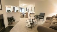 This single-floor residence showcases an open-concept design that connects the living and dining areas in a seamless, flowing layout. The living area is anchored by plush neutral carpeting and features a glass-topped oval coffee table with a wrought-iron base, complemented by a charcoal accent chair and a sofa. Tall decorative candle stands add dramatic vertical interest to the space, while potted greenery introduces natural touches throughout.

The dining area, visible from the living room, is furnished with a round table surrounded by upholstered slipcovered chairs, creating an inviting setting for gatherings. Decorative wall art and a classic table lamp add warmth and character to the adjacent console area, where a framed mirror reflects light and enhances the sense of depth. A pass-through window connects the dining space to what appears to be a kitchen, improving both functionality and interaction between rooms.

Throughout the space, a soft, warm color palette of creams and beiges creates a cohesive, tranquil ambiance. The overall aesthetic blends traditional and transitional design elements, resulting in an elegant yet comfortable living environment.