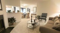 This single-floor residence showcases an open-concept design that connects the living and dining areas in a seamless, flowing layout. The living area is anchored by plush neutral carpeting and features a glass-topped oval coffee table with a wrought-iron base, complemented by a charcoal accent chair and a sofa. Tall decorative candle stands add dramatic vertical interest to the space, while potted greenery introduces natural touches throughout.

The dining area, visible from the living room, is furnished with a round table surrounded by upholstered slipcovered chairs, creating an inviting setting for gatherings. Decorative wall art and a classic table lamp add warmth and character to the adjacent console area, where a framed mirror reflects light and enhances the sense of depth. A pass-through window connects the dining space to what appears to be a kitchen, improving both functionality and interaction between rooms.

Throughout the space, a soft, warm color palette of creams and beiges creates a cohesive, tranquil ambiance. The overall aesthetic blends traditional and transitional design elements, resulting in an elegant yet comfortable living environment.