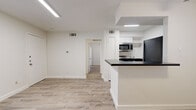 Located at 2707 Shelby Ave in Dallas, TX 75219, this single-floor, one-bedroom, one-bathroom unit offers a well-designed layout that maximizes every square foot. The open-concept living and kitchen area is a standout feature, anchored by a peninsula with dark countertops and white cabinetry, complemented by stainless steel appliances including a microwave and gas range. A distinctive white-painted exposed brick accent wall adds texture and character to the living space, while light wood-look luxury vinyl flooring flows seamlessly throughout the entire unit. Natural light enters through horizontal-blind-fitted windows, brightening the clean, neutral interior. Ceiling fans are present in both the living area and bedroom, enhancing comfort year-round. The bedroom is a comfortable, well-proportioned room with its own window, ceiling fan, and direct access to a dedicated closet. The bathroom features a full tub-shower combo with white subway tile surround and brushed bronze hardware, maintaining a crisp, clean aesthetic. A hallway connects the main living areas to the private quarters. The overall design is modern and streamlined, with a consistent white and neutral palette that creates a bright, cohesive atmosphere throughout the home.