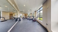 Located at Cauley Residences in Miami, FL, this fitness center offers a thoughtfully designed workout space for residents. The room is finished with durable speckled rubber flooring, a drop-panel ceiling with recessed lighting, and neutral warm-toned walls that create a clean, professional atmosphere. Large black-framed windows wrap much of the perimeter, flooding the space with natural light and providing lush, green outdoor views of the surrounding South Florida landscape — making workouts feel open and energizing. Full-length mirrors line one wall, visually expanding the space and serving a functional purpose for form monitoring during exercise. The facility is equipped with a comprehensive selection of cardio machines, including treadmills, an elliptical trainer, a stationary cycle, and a rowing machine. A dedicated strength training area features a dumbbell rack with a full range of free weights and an adjustable workout bench. The space is climate-controlled via ceiling-mounted HVAC vents. Two access doors are positioned at opposite ends of the room, ensuring convenient entry and exit. The Cauley Residences branding is prominently displayed on the walls, reinforcing the facility's identity as part of the broader residential community.