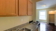 This single-floor residence offers a practical and comfortable layout designed for everyday living. The kitchen is a notable feature, outfitted with warm-toned wood cabinetry that provides generous upper storage space. The countertops feature a granite-look surface, offering durability and a classic aesthetic. A stainless steel sink sits conveniently along the counter, complemented by a gas range cooktop, making the space well-equipped for home cooking. The kitchen walls are finished in a neutral, soft yellow tone, contributing to a bright and welcoming atmosphere. A window in the adjacent room allows natural light to filter through, brightening the overall space and creating a sense of openness between areas. A traditional-style radiator is visible near the window, indicating a classic heating system consistent with established residential construction. The single-floor layout ensures easy accessibility and a seamless flow throughout the home. Overall, the space presents a straightforward and functional living environment with practical finishes and a warm, understated character well-suited for comfortable day-to-day living.