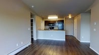 This single-floor residence offers a thoughtfully designed open-concept layout that seamlessly connects the living and kitchen areas. The space is anchored by rich, dark hardwood flooring that runs throughout, lending warmth and a sense of continuity to the interior.

The kitchen is a standout feature, equipped with dark espresso cabinetry, black stainless steel appliances including a refrigerator and microwave, and a light-toned granite or stone countertop. A peninsula island with a built-in sink serves as both a functional workspace and a natural divider between the kitchen and the open living area, making it ideal for casual dining or entertaining.

Pendant lighting hangs above the peninsula, complementing the recessed and flush-mount ceiling fixtures that provide ample illumination throughout. The neutral beige wall tones create a clean, versatile backdrop that pairs well with the darker flooring and cabinetry.

To one side of the living area, a built-in shelving unit offers organized storage, while a baseboard heating unit along the exterior wall speaks to the practical comforts of the home. Additional doorways lead to further rooms, suggesting a well-connected and functional floor plan. Overall, the space presents a modern, move-in-ready feel with a balanced blend of style and practicality.