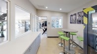 This single-floor internet café is a thoughtfully designed amenity space that combines convenience and comfort in a clean, modern layout. The interior features neutral gray and white walls with recessed lighting that creates a bright, welcoming atmosphere throughout.

A dedicated vending area lines one side of the space, equipped with both a beverage refrigerator stocked with a wide variety of drinks and a snack vending machine, ensuring guests have easy access to refreshments. A built-in white countertop with gray lower cabinetry runs along the wall, offering ample surface space for personal items or light tasks.

An interior window connects this space to the adjoining fitness center, visible through the glazed opening and accessible via a doorway, fostering a sense of connectivity between the two amenity areas. The fitness center, glimpsed through the pass-through, features exercise equipment including stationary bikes and cardio machines.

A casual seating nook provides a relaxed spot to unwind, furnished with vibrant green bar-height stools and a round table. A multi-panel black-and-white bicycle-themed art piece adorns the wall above, adding a stylish, urban touch. A window in this corner looks out to the exterior parking area, bringing in natural light. The space is finished with light speckled flooring and dark base trim throughout, giving it a polished, cohesive look.