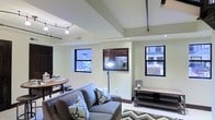 This two-floor urban residence offers a well-designed layout that balances comfort and functionality. The main living area is an inviting, open space anchored by a plush gray sofa accented with patterned throw pillows, resting atop a bold chevron-patterned area rug. A rustic media console adds warmth and contrast against the neutral, light-toned walls.

The kitchen is partially open to the living area through a pass-through window, providing a connected feel between spaces. Dark cabinetry and stainless steel appliances are visible through the opening, offering a classic and practical kitchen aesthetic.

Natural light filters into the space through multiple black-framed windows that frame views of the surrounding urban streetscape, lending a contemporary, industrial character to the interior. Recessed lighting and track lighting mounted on the ceiling provide versatile illumination throughout the day and evening.

A dining area with a round wooden table and coordinating chairs is positioned near the windows, creating an intimate setting for meals. Exposed piping along the ceiling adds an industrial loft-like touch, complementing the overall urban aesthetic. The two-floor layout provides additional living or sleeping space, making this residence both spacious and adaptable.