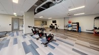 Located at 115 Wilcox St in Castle Rock, CO, this single-level fitness center is thoughtfully designed to accommodate a wide range of workout styles and training needs. The space is organized into distinct functional zones, each finished with durable flooring and illuminated by recessed panel lighting throughout.

The main cardio floor is a spacious, open area featuring treadmills, stationary bikes, and rowing machines arranged to maximize movement and flow. Large windows along the perimeter allow natural light to fill the room while providing views of the surrounding neighborhood. A strength training section includes cable machines, a full dumbbell rack, and weight benches, all positioned alongside floor-to-ceiling mirrors that enhance the sense of space.

A dedicated cycling zone houses multiple spin bikes and a mounted flat-screen television, with storage cubbies and additional strength accessories, including resistance bands, stability balls, and a cable-pulley system nearby.

The standout feature is a private studio separated by a sleek glass partition wall. This room boasts warm hardwood-style flooring, a wall-mounted ballet barre, a stall bar, large mirrors, and a dramatic floor-to-ceiling landscape mural depicting a yoga figure atop a mountain—setting a motivating, tranquil atmosphere ideal for yoga, barre, or group fitness classes.