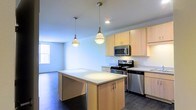 This single-floor space showcases a thoughtfully designed open-concept layout that seamlessly connects the kitchen and living areas, creating a sense of spaciousness and ease of movement throughout. The kitchen is a standout feature, equipped with a full suite of stainless steel appliances including a gas range, built-in microwave, and dishwasher. Light wood cabinetry lines the walls, offering generous storage, while clean white countertops provide ample prep space. A central kitchen island anchors the room, topped with a bright white surface and accented by two stylish pendant lights that hang overhead, adding a modern decorative touch. Dark hardwood flooring runs throughout, providing a rich contrast to the light cabinetry and neutral walls. The open living area beyond the kitchen benefits from natural light filtering in through a window, contributing to the airy and welcoming ambiance of the space. The overall aesthetic is clean, contemporary, and functional, making it an inviting environment suited for both everyday living and entertaining.