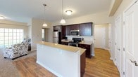 This single-floor residence offers a thoughtfully designed layout that balances comfort and functionality across its key living spaces. The kitchen serves as a central highlight, featuring rich dark cabinetry, granite-style countertops, and a full suite of stainless steel appliances including a refrigerator, range, and microwave. A large peninsula island with a clean white finish provides additional prep space and casual seating, while pendant lighting overhead adds a polished touch. Warm hardwood-style flooring runs throughout the kitchen and flows seamlessly into the adjacent living and dining areas, which are finished with plush carpet. The dining nook is brightened by a large window with views to the outdoors, and the living area offers generous wall space ideal for entertaining. The primary bedroom is carpeted and well-sized, connecting directly to a private bathroom equipped with a dual-sink vanity topped with granite-style counters. A second bathroom features a full tub-shower combination with tile flooring. Additional bedrooms are carpeted, filled with natural light from large windows, and offer ample space for various uses. Neutral warm tones throughout the space create a cohesive, inviting atmosphere.