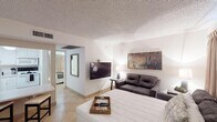 Located at 1251 N 76th St in Scottsdale, Arizona, this single-story unit offers a thoughtfully arranged layout designed for comfortable everyday living. Light-toned tile flooring flows throughout the space, complementing the crisp white walls and textured ceilings that lend a consistent, clean aesthetic from room to room.

The open living area serves as the heart of the unit, featuring a wall-mounted flat-screen TV, a dark leather loveseat, and framed artwork that adds a touch of warmth to the neutral palette. This space connects seamlessly to a dedicated dining area furnished with a dark wood table and seating for multiple guests, positioned adjacent to the kitchen.

The kitchen is well-equipped with white cabinetry, a full-size refrigerator, dishwasher, and ample counter space, making it fully functional for daily meal preparation. A decorative octagonal mirror accents the kitchen wall, adding visual interest to the utilitarian space.

The private bedroom provides a quiet retreat, complete with a ceiling fan, wall-mounted sconce lighting, bold abstract artwork, and floor-length curtains. A walk-in closet offers generous storage, and a wall-mounted TV adds convenience.

The bathroom features an extended vanity with a wide mirror, white cabinetry, tile flooring, and a shower-tub combination, finished with neatly arranged towels and tasteful framed wall art.