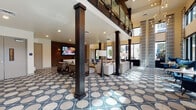 This expansive two-story amenity space is defined by its bold geometric tile flooring—a striking navy and cream circular pattern that flows seamlessly throughout both levels, creating a cohesive and visually dynamic foundation. The ground floor opens into a grand lounge area with soaring double-height ceilings, floor-to-ceiling patterned drapery, and an abundance of natural light streaming through large windows and glass doors. Dark wood columns punctuate the open space, adding architectural contrast against the neutral walls and furnishings. A generously appointed seating lounge features plush sectional sofas, layered rugs, and accent chairs arranged around a central coffee table, with views to the outdoor pool and palm-lined surroundings. A dedicated dining and wet bar area showcases a mosaic tile feature wall, large-format TV, wood communal table, and built-in cabinetry with countertops. A coffee station and staircase with glass and metal railings provide access to the second floor, which hosts additional recreational areas including a billiards table, shuffleboard, lounge seating, and a large projection screen. Exposed dark beams accent the upper level's ceiling, reinforcing the space's modern yet warm aesthetic throughout.