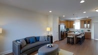 This three-floor residence is designed with an open-concept layout that connects the living, dining, and kitchen areas into one cohesive, functional space. The main living area features warm hardwood-style flooring that extends throughout, creating a sense of continuity and flow. Recessed lighting in the ceiling provides even illumination across the space, complemented by a stylish pendant light that anchors the kitchen zone.

The kitchen is equipped with stainless steel appliances, including a French door refrigerator and a gas range, and is finished with warm honey-toned cabinetry that extends to the upper walls for ample storage. A kitchen island with a light stone countertop doubles as a breakfast bar, with barstool seating providing a casual dining option. A window above the sink allows natural light to filter into the kitchen area.

The adjacent living area offers generous space for comfortable seating arrangements, with smooth, neutral-toned walls that lend a clean, contemporary aesthetic throughout. The multi-floor layout provides additional living, sleeping, or flexible-use spaces beyond the main level, making this a well-rounded and versatile residence.