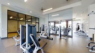 This fitness center offers a thoughtfully designed workout environment with a clean, contemporary aesthetic. The space is outfitted with a comprehensive selection of professional-grade strength training machines, strategically arranged to allow comfortable movement between equipment. The flooring features durable, rubberized material suited for high-traffic gym use, providing both safety and practicality.

Ceiling height is notably generous, contributing to an open and airy atmosphere that prevents the space from feeling cramped even when fully equipped. Overhead, linear fluorescent lighting supplements the ample natural light that filters in through large windows and glass doors along one wall, keeping the environment bright and energizing throughout the day.

A full-length mirror wall along one side of the space visually expands the room while allowing users to monitor their form during workouts. An adjacent glass-partitioned room is visible, suggesting a dedicated area for additional training activities. The neutral color palette of whites and grays, combined with the polished finishes, gives the facility a modern, professional look consistent with a well-maintained, amenity-focused fitness space.