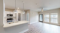 This single-floor residence on Johns Island, SC offers a thoughtfully designed open-concept layout with modern finishes throughout. Light hardwood-style flooring flows seamlessly across the living, dining, and kitchen areas, creating a cohesive and airy atmosphere. The kitchen is a standout feature, equipped with white shaker cabinetry, quartz countertops, stainless steel appliances including a refrigerator, range, microwave, and dishwasher, and a large center island with an undermount sink. Stylish pendant lighting hangs above the island, complementing the recessed lighting and built-in wine rack detail above the refrigerator. The open living area extends toward a glass door and windows fitted with blinds, allowing natural light to fill the space, while a ceiling fan adds comfort. A built-in shelving unit provides additional storage and display space. The bedrooms are generously sized, each with ceiling fans, large windows, and the same warm hardwood-style flooring. The primary bathroom features a double-sink vanity with quartz countertops, white cabinetry, black hardware accents, and a walk-in shower. The secondary bathroom includes a full soaking tub with large-format tile surround. The overall palette of soft neutral tones creates a clean, contemporary ambiance throughout.