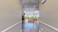 Located at 465 Buckland Hills Drive in Manchester, Connecticut, this single-story community space is designed with an open, airy layout that emphasizes natural light and architectural detail throughout. Entry is marked by a slate tile floor transitioning into warm hardwood, offering a clear visual distinction between zones as you move deeper into the space.

The main gathering area commands attention with dramatically high ceilings that rise well above standard height, creating a sense of volume and openness. A prominent stone fireplace with a rustic wood mantel serves as the focal point of the room, complemented by a wall-mounted flat-screen television above it. The surrounding walls are fitted with an array of large, grid-paned windows positioned at multiple levels — including clerestory windows near the ceiling — that flood the interior with natural light and provide views of the outdoor surroundings, including a visible pool area.

Seating is arranged to encourage both relaxed lounging and casual gathering, with cream-toned sofas and accent chairs alongside tall bistro-style tables and wood-and-chrome bar stools. A kitchen area includes dark cabinetry with stainless steel appliances, including a wall oven and refrigerator. The overall ambiance is bright, welcoming, and well-appointed.