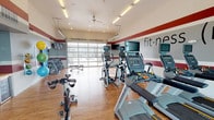 This modern fitness center is designed to support a comprehensive workout experience within a bright, open single-floor layout. The interior is distinguished by bold crimson and gray horizontal striping along the walls, warm hardwood-style flooring, and soaring ceilings fitted with a ceiling fan and recessed and overhead fluorescent lighting. A standout architectural feature is the full-height glass garage door wall that opens the space to the outdoors, allowing abundant natural light to pour in and offering views of the surrounding exterior landscape. A large floor-to-ceiling glass window panel on the opposite end further enhances the airy, open atmosphere. The main workout area is outfitted with a robust selection of Matrix-brand cardio equipment, including treadmills, ellipticals, and stationary cycling bikes, each equipped with individual display screens. A dedicated free weights zone houses a full rack of dumbbells and kettlebells alongside an adjustable weight bench, while wall-mounted medicine balls and a stability ball rack provide functional training options. A compact welcome nook near the entrance features a wooden countertop, built-in shelving, and a small beverage refrigerator stocked with water bottles, offering a convenient refreshment station. The space is branded with "fit·ness" typography across the feature wall, reinforcing its purpose-driven design.
