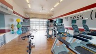 This modern fitness center is designed to support a comprehensive workout experience within a bright, open single-floor layout. The interior is distinguished by bold crimson and gray horizontal striping along the walls, warm hardwood-style flooring, and soaring ceilings fitted with a ceiling fan and recessed and overhead fluorescent lighting. A standout architectural feature is the full-height glass garage door wall that opens the space to the outdoors, allowing abundant natural light to pour in and offering views of the surrounding exterior landscape. A large floor-to-ceiling glass window panel on the opposite end further enhances the airy, open atmosphere. The main workout area is outfitted with a robust selection of Matrix-brand cardio equipment, including treadmills, ellipticals, and stationary cycling bikes, each equipped with individual display screens. A dedicated free weights zone houses a full rack of dumbbells and kettlebells alongside an adjustable weight bench, while wall-mounted medicine balls and a stability ball rack provide functional training options. A compact welcome nook near the entrance features a wooden countertop, built-in shelving, and a small beverage refrigerator stocked with water bottles, offering a convenient refreshment station. The space is branded with "fit·ness" typography across the feature wall, reinforcing its purpose-driven design.