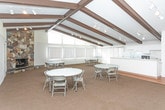 Located on Rampart Circle in Southfield, Michigan, this single-floor gathering space offers a versatile, open-plan layout well-suited for meetings, events, and community functions. The most striking architectural feature is the vaulted ceiling with prominent dark wood exposed beams running its length, lending warmth and character to the otherwise clean, white interior. Track lighting mounted along the beams ensures consistent, adjustable illumination throughout the space.

The main hall is expansive and open, with a combination of neutral carpet and hardwood flooring that defines different functional zones. A built-in counter and cabinetry area serves as a convenient service station or refreshment bar along one wall. Multiple round and rectangular folding tables with chairs are arranged throughout, allowing for flexible configurations depending on the event.

A bank of large windows fitted with vertical blinds lines one side of the hall, admitting generous natural light while maintaining privacy. A distinctive stone fireplace accent wall adds a rustic, inviting focal point to the space. An entry corridor with louvered closet doors and wood flooring provides a welcoming transition into the main area. Additional doors suggest access to utility or storage rooms, further enhancing the space's functionality.