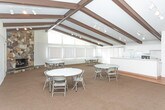 Located on Rampart Circle in Southfield, Michigan, this single-floor gathering space offers a versatile, open-plan layout well-suited for meetings, events, and community functions. The most striking architectural feature is the vaulted ceiling with prominent dark wood exposed beams running its length, lending warmth and character to the otherwise clean, white interior. Track lighting mounted along the beams ensures consistent, adjustable illumination throughout the space.

The main hall is expansive and open, with a combination of neutral carpet and hardwood flooring that defines different functional zones. A built-in counter and cabinetry area serves as a convenient service station or refreshment bar along one wall. Multiple round and rectangular folding tables with chairs are arranged throughout, allowing for flexible configurations depending on the event.

A bank of large windows fitted with vertical blinds lines one side of the hall, admitting generous natural light while maintaining privacy. A distinctive stone fireplace accent wall adds a rustic, inviting focal point to the space. An entry corridor with louvered closet doors and wood flooring provides a welcoming transition into the main area. Additional doors suggest access to utility or storage rooms, further enhancing the space's functionality.