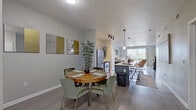 This well-appointed single-floor residence offers a thoughtfully designed open-concept layout that seamlessly connects the kitchen, dining, and living areas. The kitchen is a standout feature, equipped with stainless steel appliances including a refrigerator, microwave, and range, complemented by sleek gray cabinetry and a large center island with a white countertop and bar seating. Pendant lighting and a track light system illuminate the space, while large sliding glass doors in the living area flood the interior with natural light and provide views of the surrounding neighborhood. The dining area accommodates a round wooden table with plush velvet seating, accented by contemporary wall art in neutral and gold tones. Wood-look flooring runs throughout the main living spaces, adding warmth and continuity. The two bedrooms are carpeted for comfort, each offering access to natural light through vertical-blind-fitted windows. One bedroom features a spacious walk-in closet with built-in shelving and hanging rods. The full bathroom is finished with large-format gray tile flooring, white cabinetry, a single vanity, and a tub-shower combination, delivering a clean and functional design throughout.