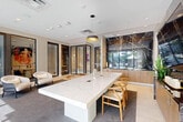 Located at 150 S Bryan Rd in Dania Beach, Florida, this elegantly appointed amenity lounge offers a refined and contemporary atmosphere designed for both productivity and relaxation. The single-floor space is thoughtfully laid out, combining a central living and meeting area with an additional flexible room, all unified by a cohesive modern aesthetic.

The main living area is anchored by a striking white marble conference-style table paired with warm wood-toned wishbone chairs, set atop a plush area rug over large-format tile flooring. A dramatic floor-to-ceiling black marble accent wall serves as a bold focal point, framing a wall-mounted flat-screen television and flanked by warm wood cabinetry. Pendant lighting above the table adds a stylish, intimate touch.

Floor-to-ceiling glass sliding doors and large windows flood the space with natural light and offer sweeping views of a lush outdoor pool terrace lined with tropical palms — bringing the South Florida landscape directly into the interior experience. Sheer drapery softens the light while maintaining the open, airy feel.

A secondary seating area features a pair of curved, upholstered lounge chairs positioned near glass-partitioned private rooms — ideal for informal meetings or focused work. Curated artwork adorns the walls throughout, adding vibrancy and personality to the space's neutral, sophisticated palette.