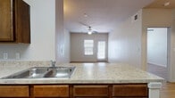 Located at 3435 E Southcross Blvd in San Antonio, Texas, this single-floor unit offers a practical and well-organized layout with consistent wood-look flooring flowing throughout every room. The open-concept kitchen serves as the heart of the space, anchored by a peninsula with a granite-style countertop and stainless steel sink. Rich dark wood cabinetry lines the kitchen walls, complemented by white appliances including a range, dishwasher, microwave, and refrigerator. A pendant light above the peninsula adds a focused decorative touch. The kitchen opens directly into the living area, which benefits from natural light streaming through a glass door and adjacent window. A ceiling fan with a light fixture keeps the living space comfortable. An arched niche adds an architectural detail near the hallway, which leads to the bedroom and bathroom. The bedroom is a well-proportioned room featuring its own ceiling fan, a window with natural light, and continued wood-look flooring. The bathroom is clean and functional, offering a full soaking tub with a tiled surround, a granite-style vanity with dark wood cabinetry, and a mirrored medicine cabinet. The unit also includes a built-in shelved closet for additional storage.