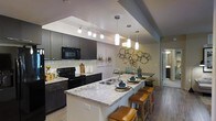 This single-floor space showcases a thoughtfully designed open-concept kitchen that serves as the centerpiece of the layout. The kitchen features dark, contemporary cabinetry paired with striking granite countertops that display elegant veining patterns in neutral tones. A large kitchen island with seating for multiple guests anchors the space, finished with matching granite and equipped with a stainless steel sink. Warm leather-upholstered bar stools line the island, adding a touch of comfort and style.

Overhead, a combination of track lighting and modern pendant lights illuminate the workspace effectively, creating a well-lit and inviting atmosphere. The classic white subway tile backsplash provides a clean contrast against the dark cabinetry, while black appliances, including a built-in microwave and range, contribute to the cohesive, modern aesthetic. Dark hardwood-style flooring runs throughout, grounding the space with warmth and continuity. Decorative wall art and metallic sculptural accents add personality to the dining area just beyond the kitchen, making the overall space feel curated and welcoming.