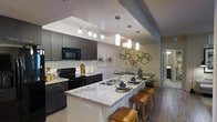 This single-floor space showcases a thoughtfully designed open-concept kitchen that serves as the centerpiece of the layout. The kitchen features dark, contemporary cabinetry paired with striking granite countertops that display elegant veining patterns in neutral tones. A large kitchen island with seating for multiple guests anchors the space, finished with matching granite and equipped with a stainless steel sink. Warm leather-upholstered bar stools line the island, adding a touch of comfort and style.

Overhead, a combination of track lighting and modern pendant lights illuminate the workspace effectively, creating a well-lit and inviting atmosphere. The classic white subway tile backsplash provides a clean contrast against the dark cabinetry, while black appliances, including a built-in microwave and range, contribute to the cohesive, modern aesthetic. Dark hardwood-style flooring runs throughout, grounding the space with warmth and continuity. Decorative wall art and metallic sculptural accents add personality to the dining area just beyond the kitchen, making the overall space feel curated and welcoming.