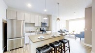 This well-appointed single-floor residence showcases a thoughtfully designed open-concept layout that seamlessly connects the kitchen, dining, and living areas into one cohesive, light-filled space. The contemporary kitchen is a standout feature, outfitted with stainless steel appliances including a refrigerator, range, and built-in microwave. Light-toned flat-front cabinetry extends to the ceiling, complemented by a subtle mosaic tile backsplash that adds texture without overwhelming the clean aesthetic. A generous kitchen island with a quartz-style countertop serves as both a prep surface and a casual dining spot, anchored by tufted black bar stools. Stylish pendant lights hang above the island, adding warmth and visual interest. The flooring throughout is a light-toned laminate or hardwood, contributing to the bright, airy feel of the space. The adjacent living area is open and inviting, with large windows that allow natural light to pour in, illuminating the neutral color palette. The overall ambiance is modern and functional, balancing clean lines with warm accents to create a comfortable and welcoming home environment.