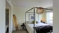 This single-floor space showcases a thoughtfully designed bedroom with a distinct urban-coastal character. At the heart of the room sits a striking four-poster bed with a sleek black metal frame accented by gold corner hardware, dressed in crisp white bedding and complemented by chartreuse and patterned accent pillows. A rich, dark fur throw adds a layer of texture and warmth. Overhead, a statement globe pendant light crafted from curved wooden slats houses an exposed Edison bulb, casting a warm and inviting glow throughout the room. A leaning wooden surfboard along one wall reinforces the laid-back coastal theme, while a black-and-white motorcycle photograph above the bed introduces an edgy, urban counterpoint. The bedside arrangement includes a geometric wire table lamp and a compact side table, adding functional elegance. Light, neutral walls and soft natural light filtering through sheer curtains keep the overall ambiance bright and airy. The combination of industrial, coastal, and bohemian design elements gives this space a curated yet relaxed personality, making it a visually compelling and comfortable retreat.