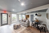 This well-appointed single-floor space is thoughtfully designed with a modern aesthetic, featuring two distinct living areas that together create a versatile and comfortable environment. Consistent wood-look flooring flows throughout, unifying the space with warmth and visual continuity. The ceilings are finished in a bold charcoal gray, adding a dramatic contrast against the crisp white walls and trim.

The primary living area is bright and welcoming, anchored by a bank of large windows dressed with patterned curtains that allow generous natural light to pour in while offering views of the surrounding greenery. A built-in nook with open shelving, a butcher block-style countertop, and a compact refrigerator serves as a convenient wet bar or refreshment station. This room also features a wall-mounted television, making it an ideal lounge or entertainment space.

The secondary living area transitions to richer, warmer wood-look flooring and serves as a casual dining and gathering zone. A round table paired with cowhide-patterned stools adds a rustic, eclectic touch, while botanical artwork and a statement pendant light lend character to the room. A gray door with a bathroom sign provides private restroom access. Together, the two areas offer a cohesive, stylishly curated space suited for both relaxation and social gatherings.