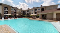 This expansive outdoor pool area offers a resort-style retreat designed for relaxation and recreation. The centerpiece is a generously sized swimming pool with clear blue water, framed by a wide surrounding deck finished in clean, light-colored pavers. The space is thoughtfully arranged to accommodate a variety of outdoor activities and lounging preferences.

Along one side of the pool, a row of chaise lounges with red cushions and large shade umbrellas provides a dedicated sunbathing and relaxation zone. On the opposite side, an outdoor dining area features multiple tables and chairs shaded by additional large umbrellas, creating an ideal setting for casual al fresco gatherings. A covered pergola or pavilion structure anchors the corner of the space, offering additional shaded seating and a defined social hub.

Lush tropical-style plantings, including palms and greenery, are woven throughout the perimeter, lending a vibrant and welcoming ambiance. The pool court is embraced on multiple sides by a multi-story building featuring a warm combination of brick and stucco exteriors, arched architectural detailing, and private balconies, creating an intimate and sheltered courtyard feel. The overall design balances elegance and comfort, making this outdoor space a standout amenity.