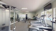 This fitness center is a thoughtfully organized single-room facility offering a comprehensive range of exercise equipment within a compact footprint. The space features a textured ceiling with crown molding trim and is finished with neutral light gray walls that create a clean, functional atmosphere. Recessed flush-mounted fluorescent light panels provide consistent overhead illumination throughout the room, while multiple windows along the perimeter walls allow natural light to filter in and offer views of the surrounding outdoor environment.

The flooring is covered in durable gray carpet, well-suited for a fitness environment. Cardio equipment is prominently featured and includes several treadmills and elliptical machines arranged along one side of the room. A multi-station cable weight machine serves as a central strength-training fixture, complemented by an adjustable workout bench, a full dumbbell rack with a range of free weights, a stability ball, and a wall-mounted television for entertainment during workouts.

A large mirrored wall expands the perceived depth of the space and is a practical feature for monitoring exercise form. A standout design element is a bold black-and-white motivational mural covering one full wall, featuring fitness-themed typography and graphics that energize the overall ambiance of the room.