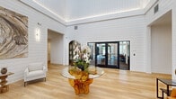 This single-floor space serves as an elegant lobby and corridor area, thoughtfully designed to make a lasting first impression. The main lobby is characterized by soaring ceilings with recessed tray lighting that casts a warm, ambient glow across the crisp white shiplap walls. Light hardwood flooring extends throughout, lending warmth and continuity to the space.

A striking centerpiece table — featuring a glass top supported by a sculptural natural wood root base — anchors the lobby, complemented by fresh orchid arrangements. Surrounding seating areas include clean-lined white and cognac leather furnishings, while large-scale nature-themed artwork, including wood grain and leaf motifs, adds organic texture to the walls. An arched display cabinet and French doors opening to an outdoor area enhance both functionality and visual depth, allowing natural light to filter inward.

The connecting hallways maintain the same light hardwood flooring and white walls, creating a seamless transition. A bold navy blue accent wall featuring a built-in water fountain station adds a distinctive design moment, while framed artwork and natural wood benches line the corridor. Recessed lighting keeps the hallways bright and welcoming, reinforcing the space's cohesive, nature-inspired aesthetic throughout.