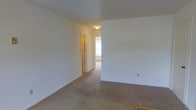 This single-floor residential space offers a clean, straightforward layout designed for comfortable everyday living. The interior is characterized by neutral white walls throughout, creating a bright and versatile backdrop that adapts easily to a variety of furnishing styles and décor preferences.

The living area features wall-to-wall carpeting in a warm beige tone, providing a soft and comfortable underfoot feel. The open layout allows for a natural flow between spaces, with a hallway connecting the main living area to additional rooms beyond. A flush-mounted ceiling light fixture provides functional overhead illumination, while a window at the end of the hallway invites natural light deep into the interior, enhancing the sense of openness.

The passage visible from the main room leads to a secondary space that also benefits from natural light, suggesting a well-considered layout that maximizes brightness throughout. A door with a classic paneled design is visible along the hallway, offering access to an additional room or utility space.

Overall, the space presents a clean, move-in-ready environment with a simple and functional design. The consistent use of neutral tones across walls, trim, and flooring contributes to a cohesive and calm ambiance throughout the unit.