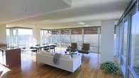 This single-floor space showcases a bold mid-century modern aesthetic, centered around an expansive wall of floor-to-ceiling wraparound windows that flood the interior with natural light and frame sweeping views of surrounding woodland. The open-plan layout connects the living and dining areas in a seamless, airy flow, enhancing the sense of spaciousness throughout.

The living area features warm hardwood flooring, a sleek white sofa paired with olive-toned lounge chairs on a patterned area rug, and a low-profile coffee table — all arranged to take full advantage of the panoramic outdoor views. A sculptural globe pendant light anchors the dining zone, where a round black table is surrounded by matching chairs, creating an elegant yet understated setting.

Architectural details elevate the space further: clean white walls and ceilings contrast with the natural wood tones of the floor and exposed exterior roof elements visible through the glass. Recessed skylights add supplemental light from above, while the wraparound glazing ensures the boundary between interior and nature feels intentionally blurred. The overall ambiance is calm, refined, and thoughtfully designed — a space that balances modernist architecture with livable warmth.