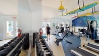 The Quail Ridge Drive Fitness Center in Plainsboro, NJ, is a well-equipped, single-floor facility designed to support a comprehensive workout routine. The space features vaulted ceilings that lend an open, airy feel, complemented by white walls and large windows that allow abundant natural light to fill the room. Views of the adjacent pool area are visible through glass-paneled doors, enhancing the bright, welcoming atmosphere.

The fitness center is divided into distinct workout zones. The cardio area houses treadmills and elliptical machines, while the strength training section includes a multi-station cable machine and an adjustable weight bench. A full rack of dumbbells in graduated sizes lines one wall, accompanied by a large mirror that visually expands the space. Additional functional training tools, including a BOSU ball, are also available.

A standout decorative element is the vibrant ocean wave mural that spans one wall, paired with surfboards mounted to the ceiling and bold yellow industrial-style pendant lighting, giving the space an energetic, beach-inspired character. The flooring combines neutral speckled tile with blue rubberized mat sections beneath the equipment. Framed artwork and clean architectural lines round out a polished, motivating environment.