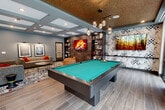 Located at 4500 University Drive in Fairfax, VA, this single-floor community lounge is a well-appointed amenity space designed for socializing and recreation. The layout is open and expansive, seamlessly blending distinct activity zones within a cohesive, modern aesthetic.

The main gathering area features a large sectional sofa and accent armchair arranged around a patterned area rug, with framed artwork illuminated by wall-mounted sconces and a flat-screen TV set against a richly tiled accent wall flanked by built-in bookshelves. An eye-catching geometric coffered ceiling with recessed lighting defines this lounge zone.

Adjacent to the seating area, a full-size pool table with teal felt commands the center of the space beneath a striking cluster pendant light fixture. A bold mosaic-style mural adds artistic character to the surrounding wall. A foosball table rounds out the game area, catering to a variety of recreational interests.

On the opposite end, a sleek kitchenette features a marble-topped island with bar seating, warm wood cabinetry, open shelving, and pendant lighting — ideal for casual entertaining. The flooring transitions between geometric patterned wood tile and wide-plank hardwood, adding visual interest throughout.

Floor-to-ceiling windows and glass sliding doors flood the space with natural light and provide direct views of and access to an outdoor courtyard, creating a bright and inviting atmosphere throughout.