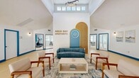 The Aqua Bay Apartments clubhouse is a thoughtfully designed community hub that blends modern aesthetics with functional amenities. The space is anchored by a dramatic vaulted ceiling that rises generously overhead, bathed in natural light streaming through a row of clerestory windows. A striking feature wall serves as the visual centerpiece, showcasing a warm wood-slatted archway in gold and blue tones, flanked by the bold Aqua Bay Apartments branding.

The lounge area is furnished with a plush blue sofa, wood-framed armchairs, and a sleek coffee table arranged over a patterned area rug, creating an inviting gathering space. Blue-trimmed French doors open to an outdoor area, further enhancing the sense of openness and connection to the exterior.

The reception area features a clean, modern desk beneath layered circular pendant lights, set against a richly upholstered deep-blue accent wall with a tufted panel design. Adjacent to reception, glass-walled enclosures provide a transparent view into a well-equipped fitness center, complete with cardio machines and strength training equipment. Throughout the space, light-toned flooring, crisp white walls, and consistent blue accent details tie the design together into a cohesive, welcoming environment.