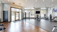 Located at 391 17th St NW in Atlanta, GA, this expansive fitness center at Farrington Apartments spans a single floor and is thoughtfully divided into two distinct exercise rooms connected by a hallway. The main strength and cardio room features rich, polished concrete floors with warm amber tones, ceiling fans, recessed lighting, and large windows that flood the space with natural light and views of the surrounding neighborhood. This area is equipped with cable machines, weight benches, a Smith machine, resistance equipment, and a row of cardio machines including treadmills, ellipticals, and stationary bikes. A vibrant cycling-themed mural serves as a bold focal point, adding an energetic artistic element to the space. Wall-mounted televisions provide entertainment throughout. The second exercise room offers a dedicated functional training studio, featuring hardwood-style flooring, floor-to-ceiling mirrors that enhance the sense of space, and natural light from large windows. This studio is outfitted with TRX suspension trainers, kettlebells, resistance bands, stability balls, a BOSU trainer, and a heavy bag — supporting a wide range of training modalities. Together, the two rooms provide a comprehensive, well-designed fitness environment suited for both strength training and dynamic workouts.