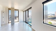 This contemporary 2-bedroom, 1-bathroom residence offers a sleek, urban living experience defined by clean lines and an industrial-modern aesthetic. Polished concrete floors run throughout the entire unit, providing a cohesive and low-maintenance foundation that complements the minimalist design. The open living room is a standout space, featuring expansive floor-to-ceiling sliding glass doors and windows that frame impressive panoramic views of the city skyline, a river, and surrounding bridges, flooding the room with natural light. A distinctive exposed concrete structural column adds an architectural accent to the living area.

Both bedrooms are well-proportioned and bright, each benefiting from large black-framed windows with elevated urban views. The primary bedroom includes a built-in wardrobe system with shelving, drawers, and hanging rails, along with a ceiling fan. The second bedroom also features a closet and convenient access to the shared bathroom.

The bathroom is finished in a clean, modern style with white subway tile in the walk-in shower, a floating vanity with an undermount sink, and a frameless mirror. Two connecting hallways provide efficient circulation between the rooms. The overall ambiance is bright, airy, and distinctly urban, ideal for those seeking a contemporary high-rise lifestyle.