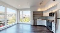 This well-appointed single-floor unit showcases a clean, modern aesthetic throughout. The open-concept living and kitchen area is a standout feature, anchored by expansive floor-to-ceiling windows that flood the space with natural light and frame views of the surrounding urban landscape, including a nearby elevated roadway and lush tree canopy. Dark hardwood-style flooring runs continuously through the main living areas, creating a warm contrast against the crisp white walls and ceilings.

The kitchen is thoughtfully designed with white cabinetry, a classic farmhouse sink, light blue-grey subway tile backsplash, warm-toned open wood shelving, and a full suite of stainless steel appliances, including a range, microwave, and dishwasher. Track lighting overhead adds a modern, functional touch.

The bedrooms are bright and simply finished, each featuring the same durable hardwood-style flooring, white walls, and windows that bring in natural light and urban views. Wall sconces provide accent lighting in the sleeping areas.

Two bathrooms serve the unit: one features a full soaking tub with a curved shower rod and a vanity with dual under-mount sinks and grey countertops, while the second offers a standalone walk-in shower with a frosted door. Both bathrooms continue the cohesive modern palette of white finishes and wood-look flooring.