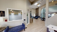 The WestEnd at Ten community clubhouse is a thoughtfully designed single-floor facility that makes an immediate impression with its dramatic vaulted ceilings and abundant natural light streaming through skylights and floor-to-ceiling glass entry doors. Light wood-look flooring flows throughout, tying together the open, airy layout.

Upon entry, a sleek reception desk with a digital check-in kiosk and a cushioned bench greet visitors, adjacent to a frosted-glass leasing office. The grand main lounge features soaring ceiling heights accentuated by a striking floor-to-ceiling reclaimed wood fireplace surround with a black marble hearth, creating a bold focal point. Comfortable seating in rich tones of navy and teal complement the vibrant abstract artwork adorning the walls.

A dedicated lounge area with a large sectional sofa, wall-mounted television, and accent lighting offers a relaxed retreat, while a separate dining area with a round wood table provides an inviting communal space. A carpeted staircase with a glass railing leads to a mezzanine level overlooking the main floor.

The community kitchen is equipped with stainless steel appliances, dark granite countertops, white cabinetry, and a breakfast bar with seating, with windows offering views of the outdoor pool area. Ceiling fans and recessed lighting throughout ensure comfort and functionality across the entire space.