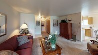 This single-floor residence at 3418 SE Navigation Ln, Unit 101 in Port Orchard, Washington offers a well-organized layout with comfortable living spaces throughout. The spacious living room serves as the centerpiece of the home, featuring a tile-surround fireplace, plush wall-to-wall carpeting, and multiple windows that allow natural light to fill the room. The living area connects seamlessly to a dining space equipped with a sliding glass door that opens to a view of a lush green outdoor landscape, making it an inviting spot for everyday meals or entertaining. The galley-style kitchen is efficiently designed with warm wood cabinetry, ample counter space, a full suite of appliances including a dishwasher and electric range, and easy-care tile flooring. The residence includes multiple bedrooms, each with carpeted floors and closet space. The primary bedroom is generously sized with a window for natural light and access to a well-appointed bathroom featuring a wide vanity, an extended countertop with a built-in dressing area, warm wood cabinetry, and a large wall mirror with overhead lighting. Additional bedrooms offer closet storage and windows for ventilation and light. The overall ambiance of the home is warm and comfortable, with neutral tones and practical finishes throughout.