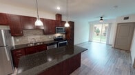 This single-floor residence offers a thoughtfully designed open-concept layout that seamlessly connects the kitchen and living areas. The kitchen is a standout feature, showcasing rich dark wood cabinetry, striking dark granite countertops, and a full suite of stainless steel appliances, including a refrigerator, range, microwave, and dishwasher. A tile backsplash adds texture and visual interest, while pendant lighting above the kitchen island provides both functional illumination and a stylish accent. The open living area extends naturally from the kitchen, bathed in natural light from large sliding glass doors that offer views of a lush green outdoor space, complemented by a ceiling fan overhead. Hardwood-style flooring runs throughout, lending a cohesive and contemporary feel to the interior. The bedroom features a tall window with views of mature trees and a well-maintained yard, creating a calm and bright retreat. The bathroom is cleanly finished with a dark wood vanity, white countertop, a tub-shower combination, and light tile flooring. Recessed lighting and neutral wall tones throughout the space contribute to a clean, modern aesthetic that is both functional and inviting.