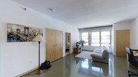 This single-floor space showcases a clean, modern aesthetic defined by its distinctive polished concrete floors, which lend a sleek, industrial character to the interior. The living area serves as the central gathering space, furnished with a light gray sofa positioned to face a television set atop a media console. A light-toned area rug anchors the seating arrangement, softening the reflective floor beneath. Large horizontal windows along one wall allow generous natural light to fill the room, enhancing the open and airy ambiance. White walls throughout keep the space feeling bright and neutral. A textured ceiling adds subtle depth overhead. Near the entrance, a set of natural wood-toned double doors provides closet storage, while a doorway leads to an adjacent room visible in the background. A floor lamp with a classic shade and a piece of cityscape wall art add decorative touches to the space. A small potted plant near the media area introduces a hint of greenery. Overall, the space balances industrial and contemporary elements in a practical, well-lit layout.