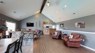 Located at Blankenbaker Crossings, this single-floor community space combines rustic charm with modern design across an open, flowing layout. Soaring vaulted ceilings with exposed wood beams define the main gathering area, which is anchored by rich hardwood-style flooring throughout. The space is divided into several functional zones that transition seamlessly into one another.

The lounge area features multiple leather armchairs with ottomans arranged around a wall-mounted flat-screen television, complemented by decorative wall art with an equestrian theme. Industrial-style pendant lighting and track lighting illuminate the space, enhancing the warm, welcoming atmosphere.

A central kitchen and service counter acts as a natural dividing element, clad in wood-toned paneling and stacked stone accents. White cabinetry and a built-in microwave are visible behind the counter, lending a clean, functional aesthetic.

The dining area accommodates a large table with mixed seating and is highlighted by a dramatic floor-to-ceiling stacked stone fireplace. A generously sized window floods the room with natural light and frames outdoor views, while wainscoting along the lower walls adds a refined architectural detail.

Overall, the space presents a cohesive rustic-modern ambiance suited for community gatherings and social events.