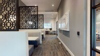 This well-appointed single-floor space combines functionality and refined design, creating a professional yet welcoming environment. The interior is anchored by a sophisticated color palette of cool grays and whites, complemented by textured, multi-toned carpeting that adds warmth underfoot throughout.

A standout feature is the striking dark lattice screen partition, which elegantly divides the lounge seating area from the business center workstations, offering a sense of privacy without sacrificing openness. The lounge area includes upholstered booth-style seating with clean-lined white tables, ideal for informal meetings or relaxed work sessions.

The business center is equipped with multiple computer workstations positioned along a built-in desk, bathed in natural light from large windows that frame outdoor greenery. Nearby, a glass-paneled door with sleek black framing leads to an adjacent corridor, reinforcing the space's modern industrial aesthetic.

A dedicated conference room, visible through floor-to-ceiling glass walls, features a large central table surrounded by contemporary rolling chairs and pendant lighting overhead, with ample natural light filtering through expansive windows.

A thoughtfully designed coffee bar area rounds out the space, showcasing a bold black-and-white geometric tile accent wall, dark wood cabinetry, and a commercial coffee machine — a functional yet stylish touch. Curated wall art adds personality throughout.