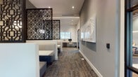 This well-appointed single-floor space combines functionality and refined design, creating a professional yet welcoming environment. The interior is anchored by a sophisticated color palette of cool grays and whites, complemented by textured, multi-toned carpeting that adds warmth underfoot throughout.

A standout feature is the striking dark lattice screen partition, which elegantly divides the lounge seating area from the business center workstations, offering a sense of privacy without sacrificing openness. The lounge area includes upholstered booth-style seating with clean-lined white tables, ideal for informal meetings or relaxed work sessions.

The business center is equipped with multiple computer workstations positioned along a built-in desk, bathed in natural light from large windows that frame outdoor greenery. Nearby, a glass-paneled door with sleek black framing leads to an adjacent corridor, reinforcing the space's modern industrial aesthetic.

A dedicated conference room, visible through floor-to-ceiling glass walls, features a large central table surrounded by contemporary rolling chairs and pendant lighting overhead, with ample natural light filtering through expansive windows.

A thoughtfully designed coffee bar area rounds out the space, showcasing a bold black-and-white geometric tile accent wall, dark wood cabinetry, and a commercial coffee machine — a functional yet stylish touch. Curated wall art adds personality throughout.