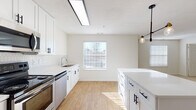 This single-floor residence offers a clean, contemporary design characterized by a bright, neutral color palette and a seamless blend of hard and soft flooring surfaces. The kitchen serves as the focal point of the home, showcasing crisp white shaker-style cabinetry with matte black hardware, white countertops, and a classic hexagonal tile backsplash. A central island provides additional prep and storage space, while stainless steel appliances — including a range, microwave, refrigerator, and dishwasher — deliver full functionality. Light wood-look flooring runs throughout the kitchen and dining area, complemented by natural light streaming in through a window fitted with blinds. A stylish multi-bulb pendant light fixture adds a modern touch above the island. The open layout connects the kitchen directly to a spacious living area, which transitions to soft, neutral carpet underfoot. A ceiling fan with a built-in light fixture keeps the living space comfortable. The bedrooms are generously proportioned, each featuring plush carpeting, ceiling fans, and ample natural light. The bathroom is finished with wood-look flooring, a vanity with under-sink storage, a wall-mounted mirror cabinet, and a tiled shower-tub combination accented with a classic black tile border. Overall, the space presents a well-maintained, move-in-ready interior with a cohesive and welcoming aesthetic.